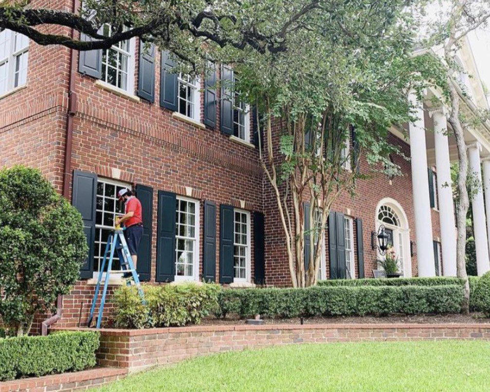 A person on a ladder cleans a window of a grand brick house with dark shutters.