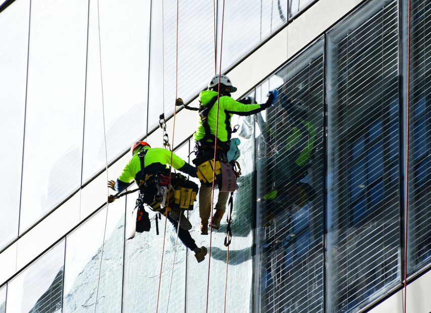Two window cleaners in safety gear are suspended by ropes, cleaning a modern high-rise building's glass facade.
