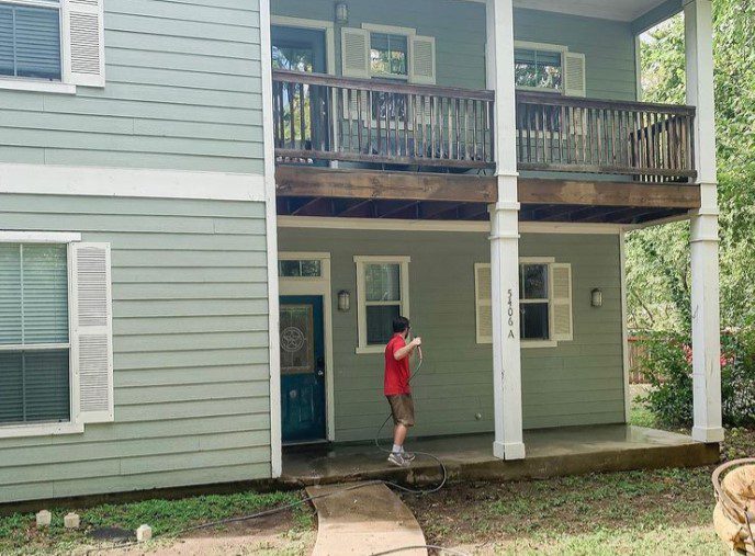 A man in a red shirt power washes the concrete porch of a two-story