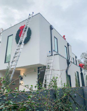 Ladders lean against a modern white house adorned with Christmas wreaths and red bows. TITLE