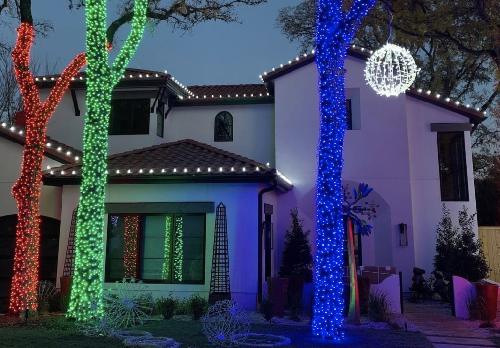 A modern white house and its trees are brightly decorated with red, green, and blue holiday lights