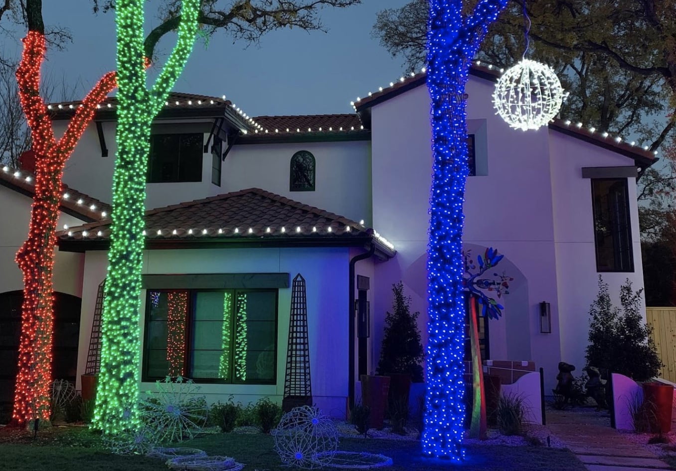 A modern white house and its trees are brightly decorated with red, green, and blue holiday lights