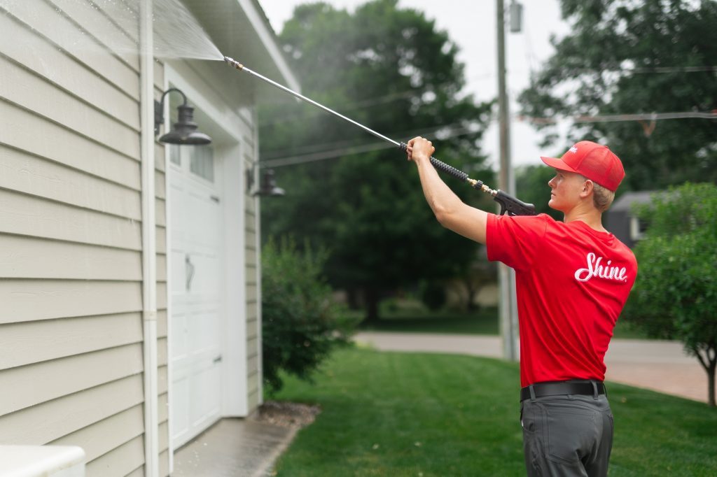 A person in a red uniform power washes the siding and eaves of a house. TITLE