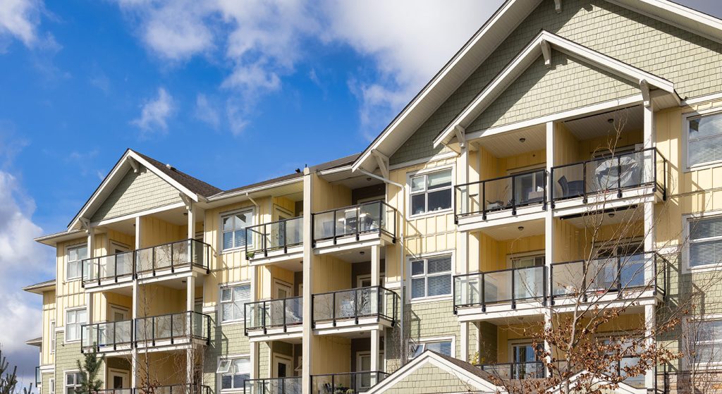 A modern multi-story condominium building with light green and yellow siding under a bright blue sky.