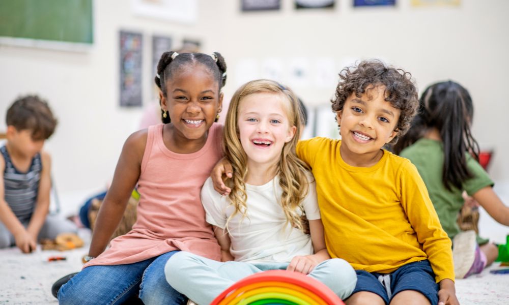 Three smiling diverse children sit together with arms around each other in a classroom. TITLE