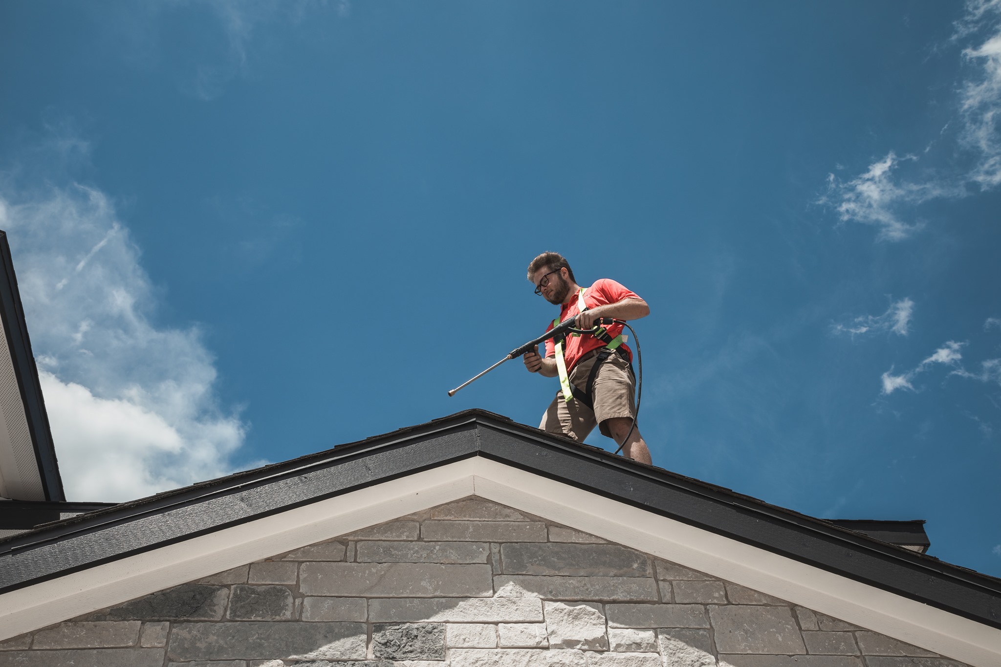 A man in a safety harness pressure washes a house roof against a blue sky.