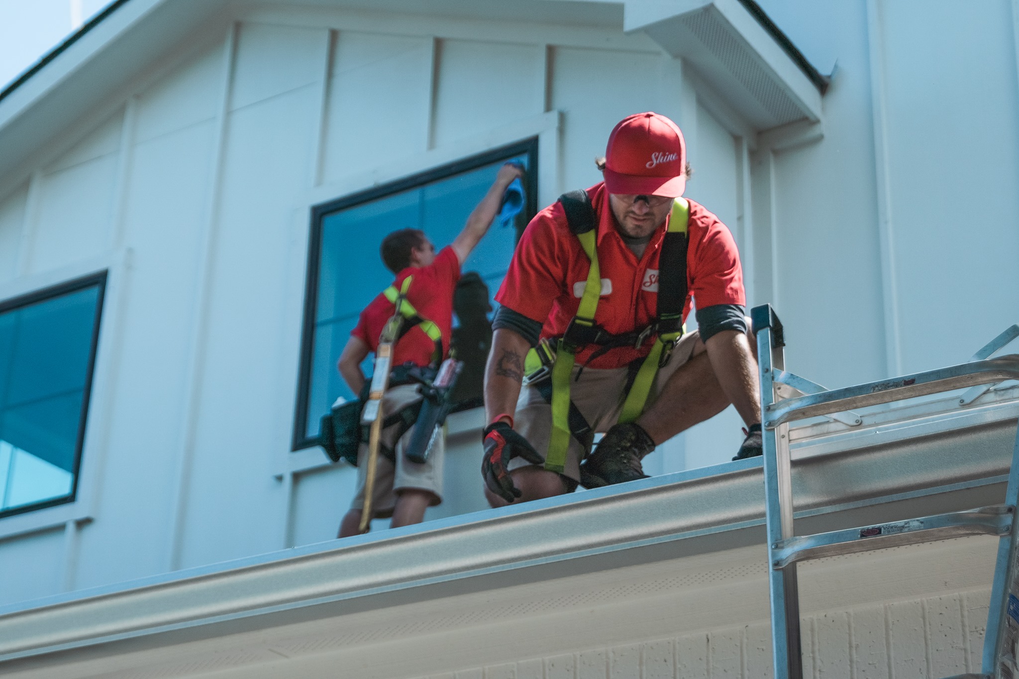 Two workers in red shirts and safety harnesses clean windows and gutters on a modern white building.