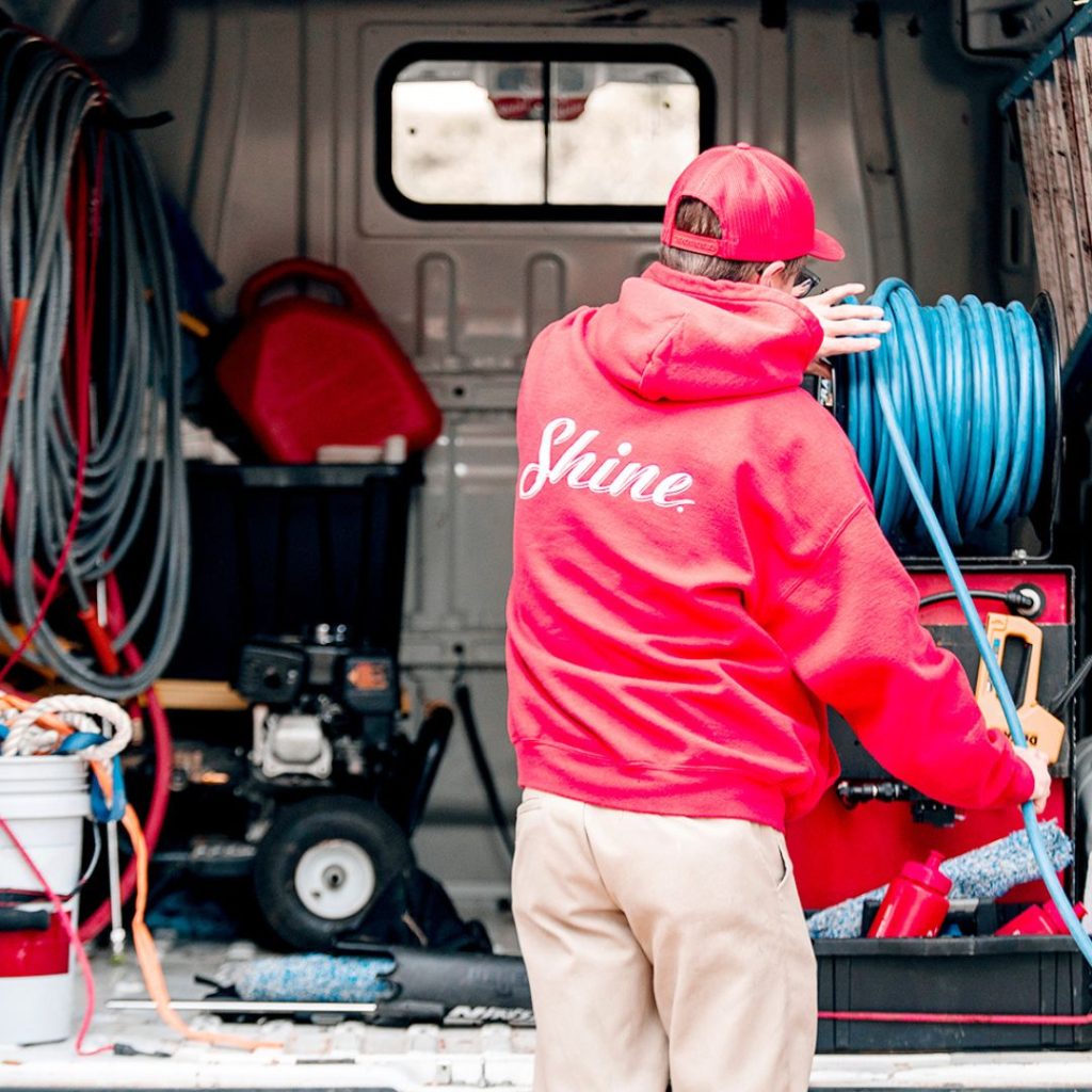 A worker in a red 'Shine' hoodie adjusts a blue hose reel inside a service