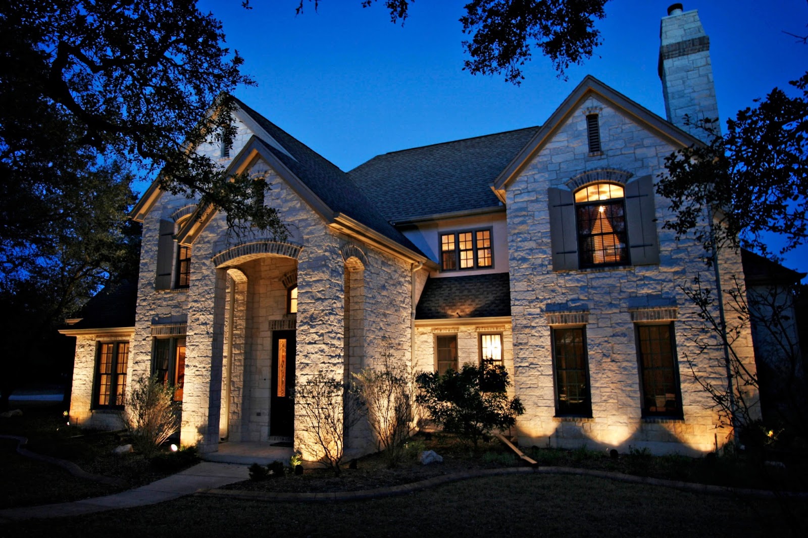 A stately stone house with an arched entry is brightly illuminated by landscape lighting at dusk.