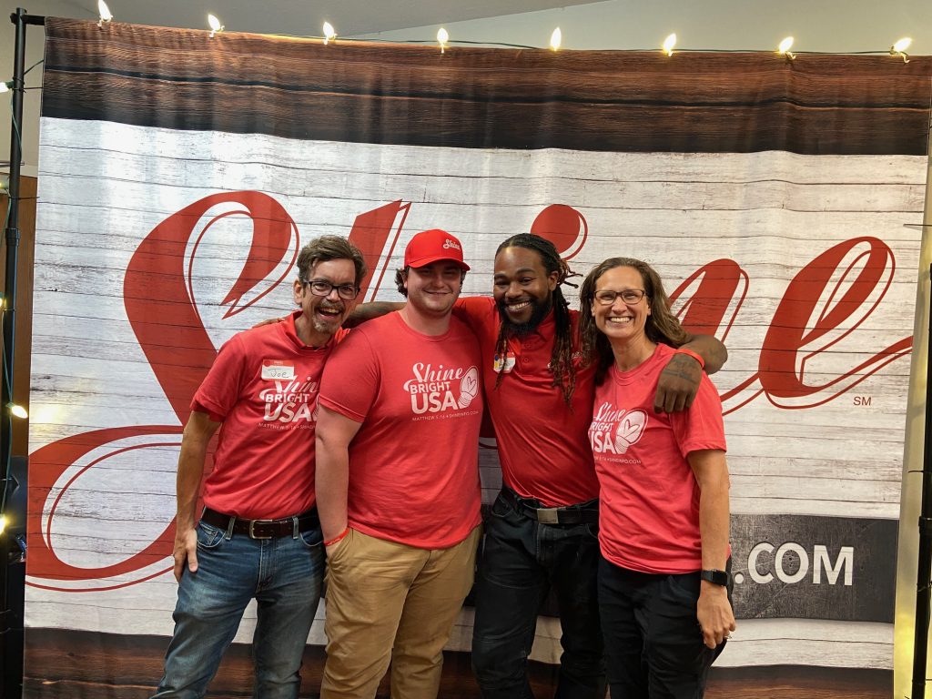 Four cheerful individuals in red "Shine Bright USA" shirts pose against a rustic wooden backdrop