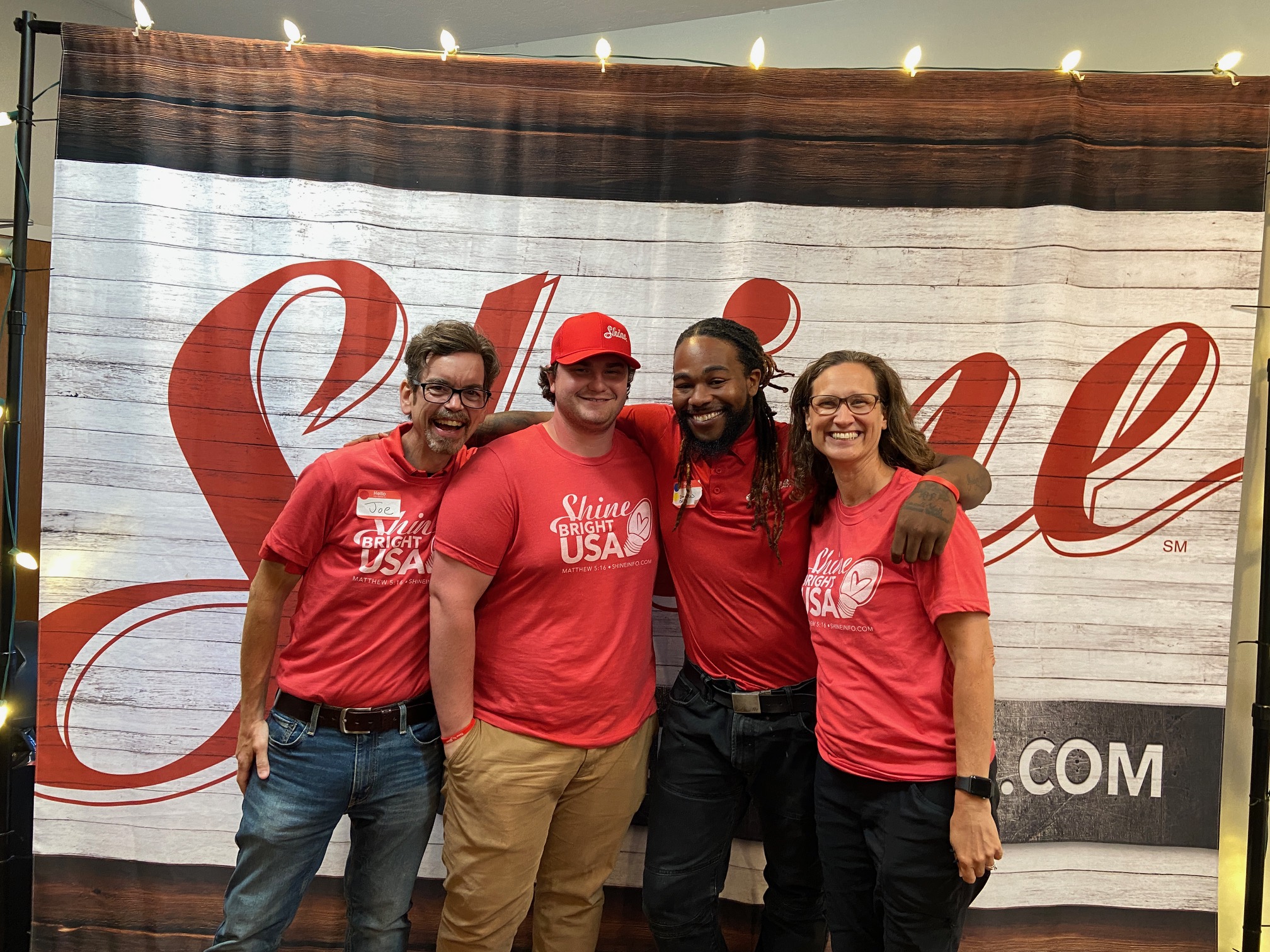 Four cheerful individuals in red "Shine Bright USA" shirts pose against a rustic wooden backdrop