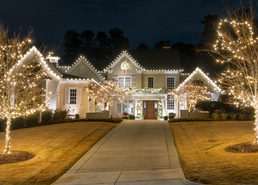 Elegant home and bare trees brightly illuminated with warm white holiday lights at night.