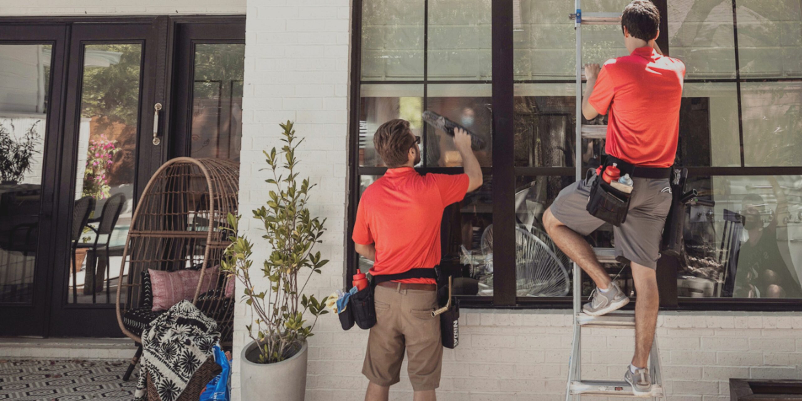 Two men wearing red shirts professionally clean the exterior windows of a modern home.