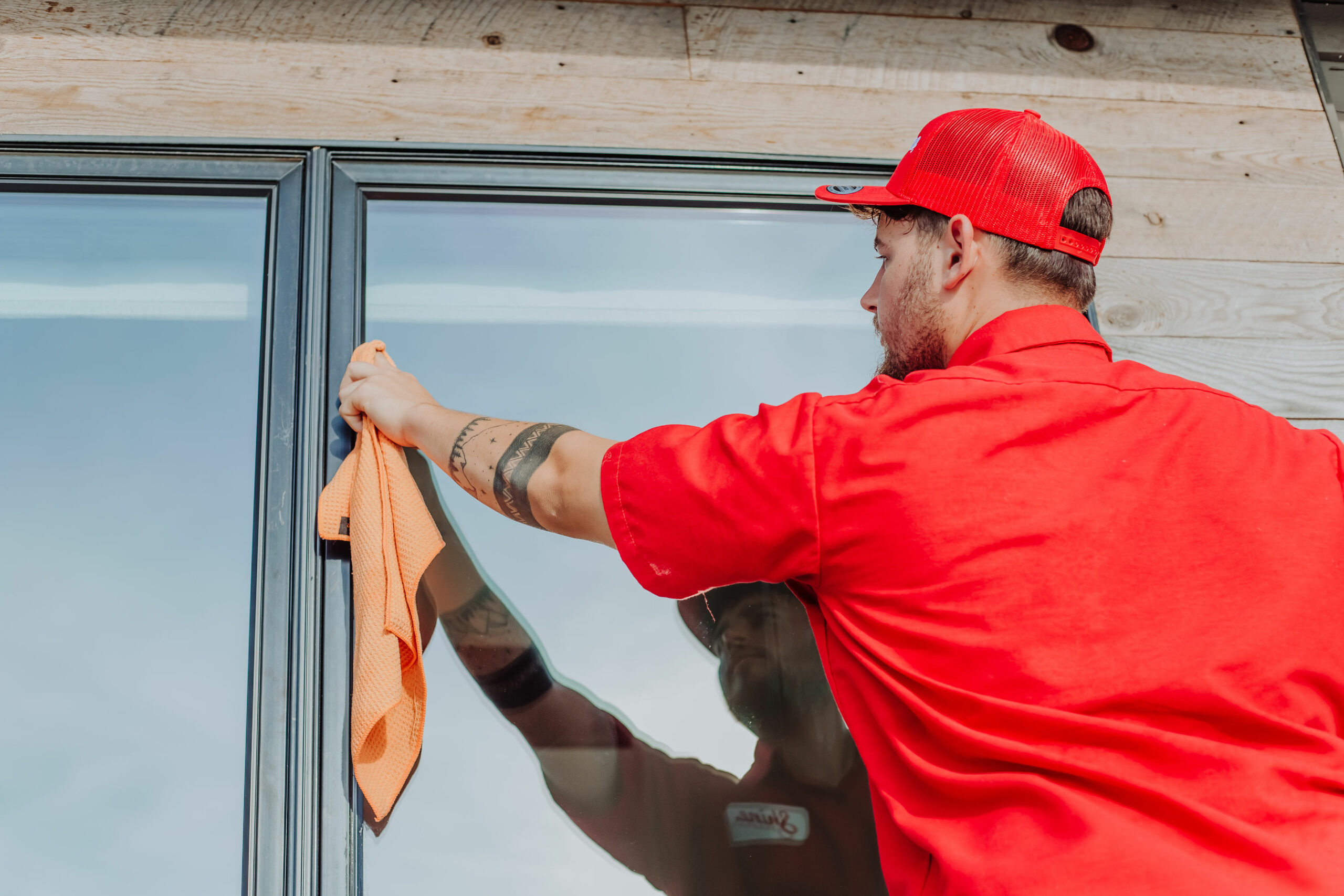 A man in a red shirt and cap meticulously cleans a large window with an orange cloth.