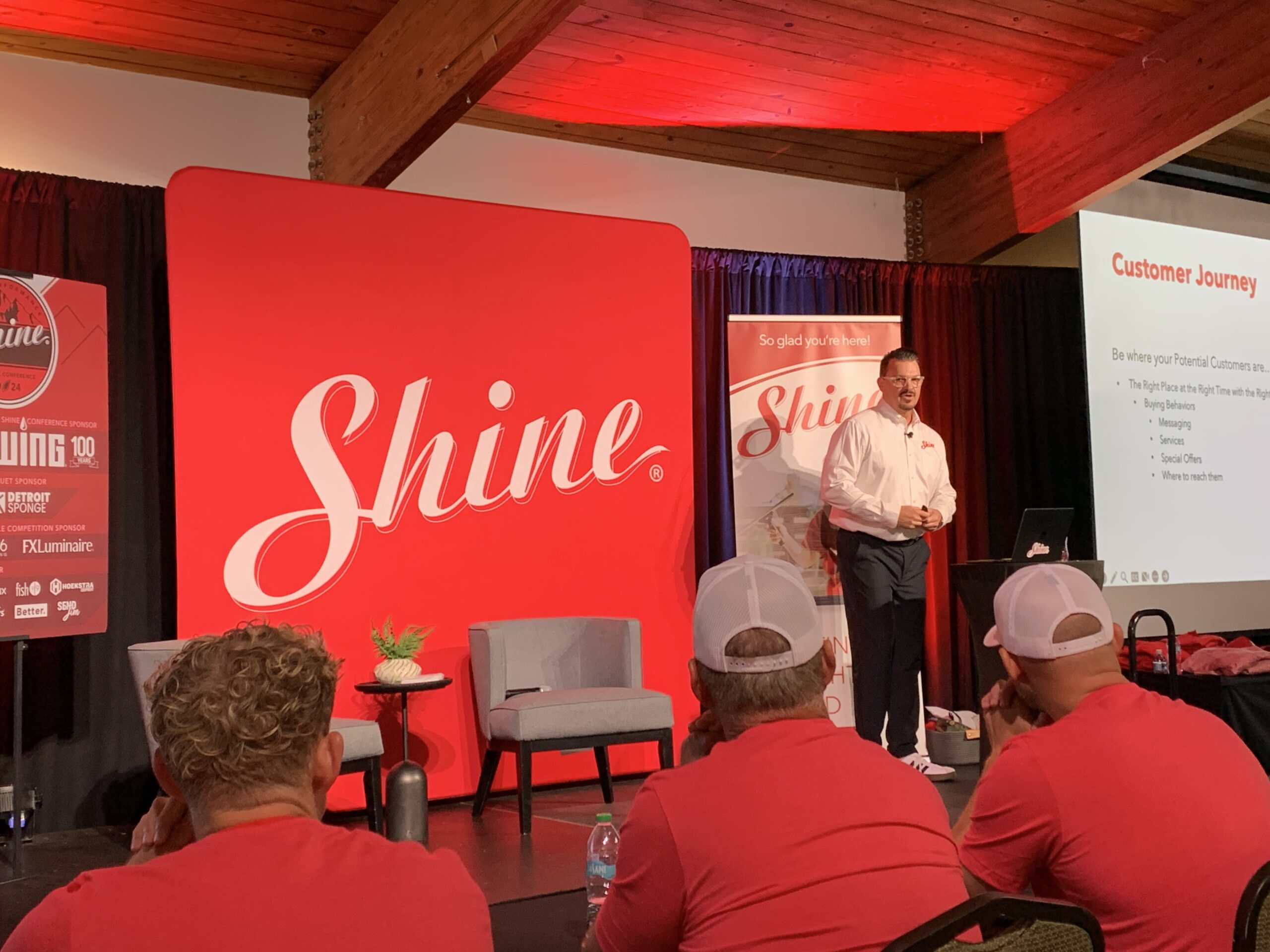 A presenter speaks to an audience in a conference room with a large red 'Shine' banner