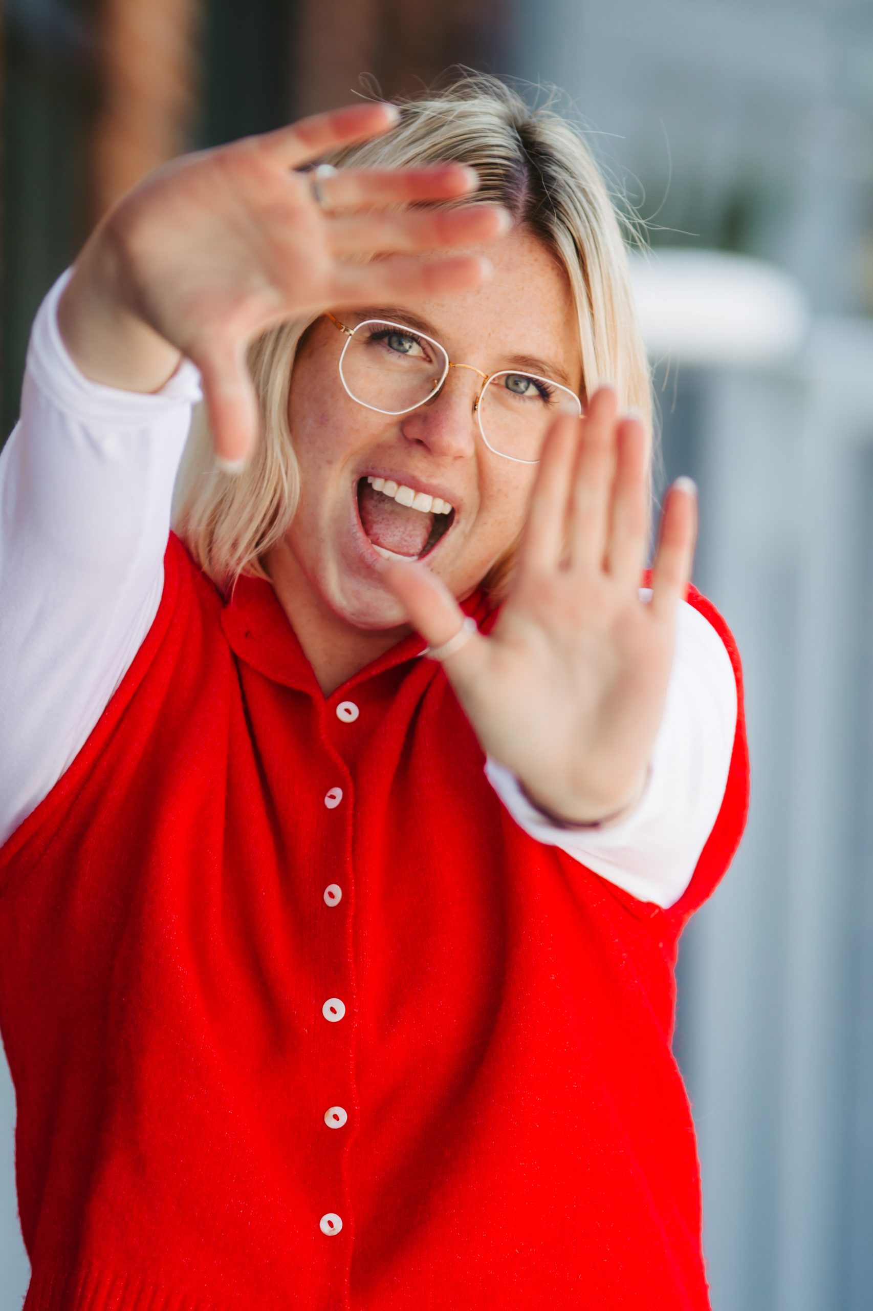 A cheerful blonde woman with glasses frames a shot with her hands, looking excited.