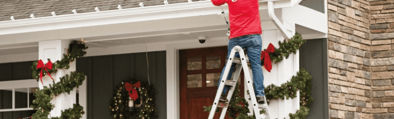 A person on a ladder hangs festive Christmas lights on a house decorated with garland and wreaths.
