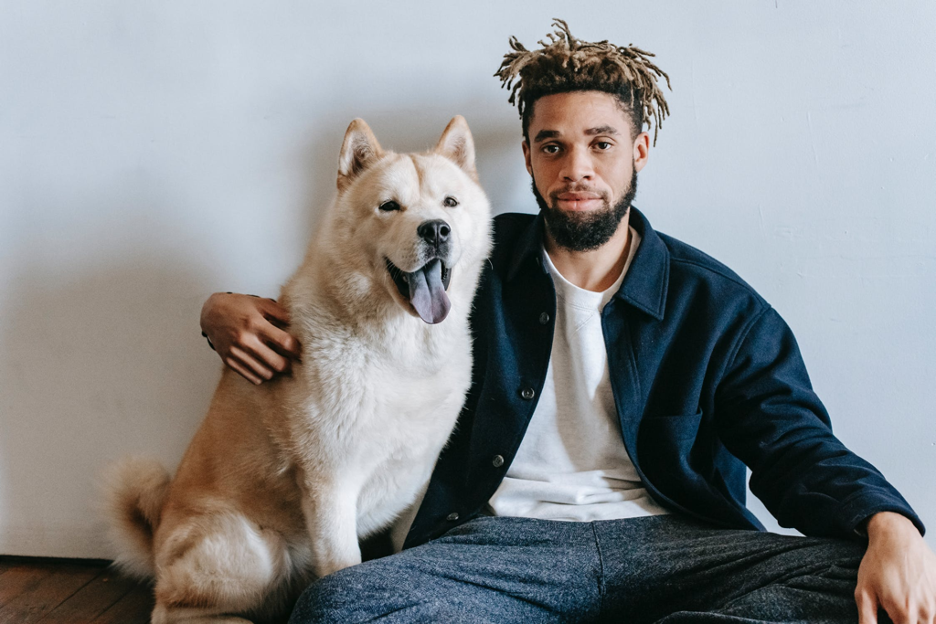 A man with dreadlocks and a beard sits with his arm around a happy cream-colored
