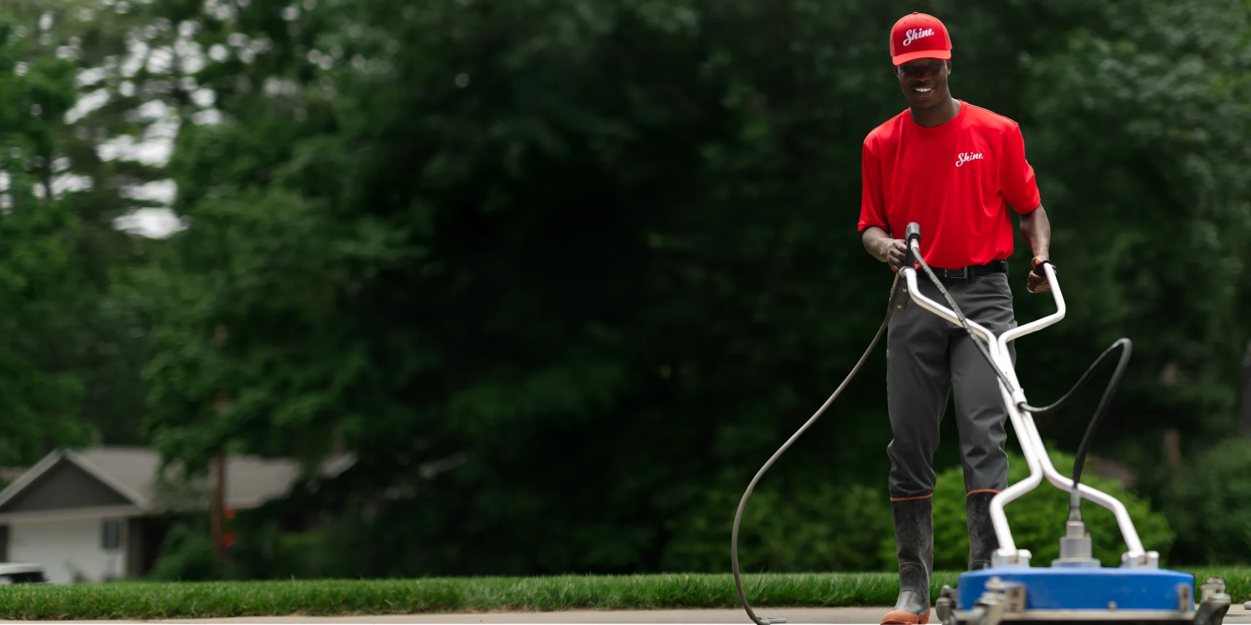A smiling man in a red "Shine" uniform pressure washes a concrete surface with