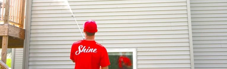 A person in a red "Shine" shirt pressure washes light-colored house siding.