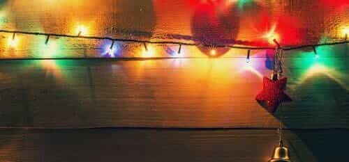Multicolored string lights illuminate a rustic wooden background with a hanging red star and bell. TITLE