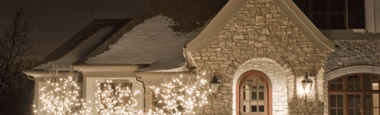 Warm white string lights illuminate a stone house with snow on its roof at night. TITLE: