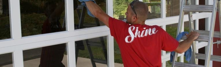 Professional window cleaner in a red "Shine" shirt wipes a large window from a ladder