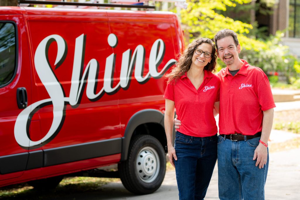 Smiling man and woman in red Shine shirts stand proudly beside a red Shine service van.