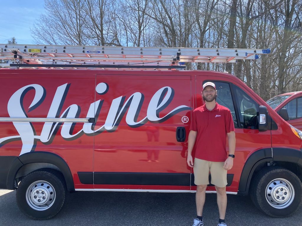 A smiling man in a red polo shirt stands beside a red Shine service van with a ladder on top.