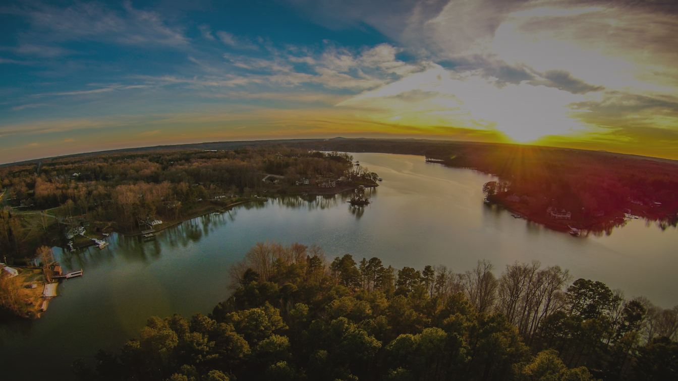 Aerial view of a wide lake surrounded by forested shores and houses under a vibrant sunset.